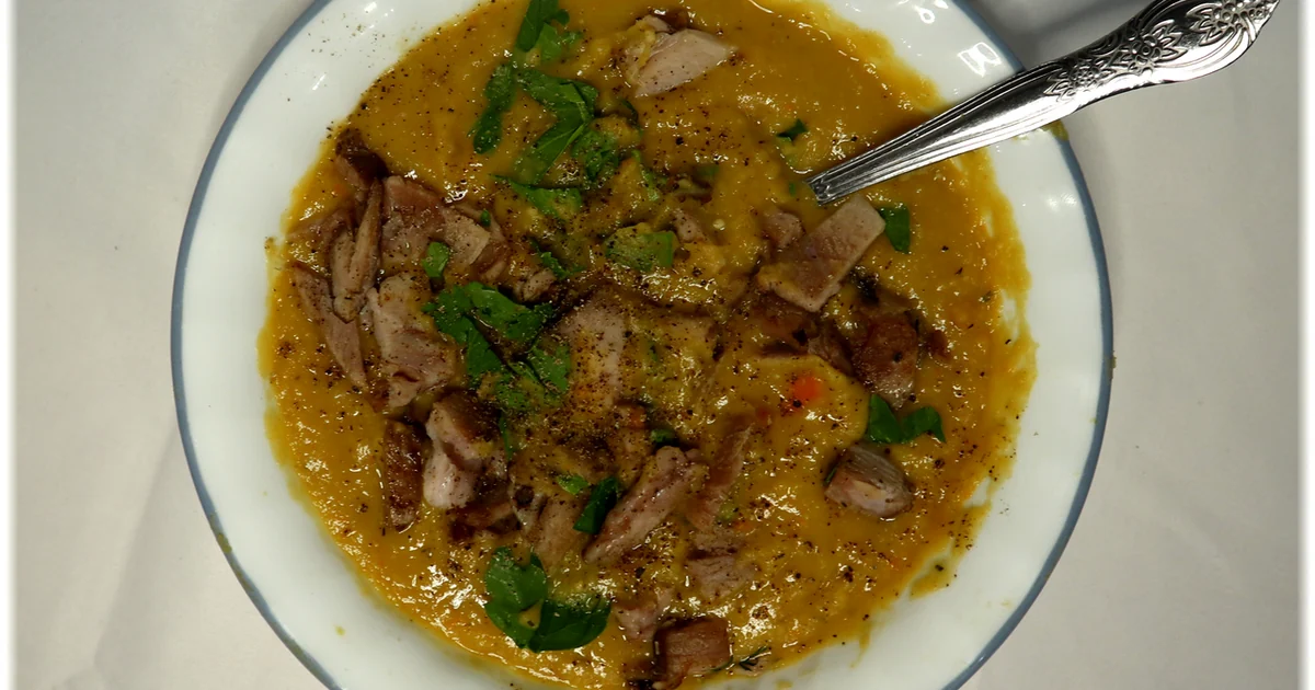 A bowl of Quebecois-style pea soup. The soup base is purred yellow split-peas and garden vegetables. Shredded ham-hock is mixed in. The soup is garnished with parsley and black pepper.