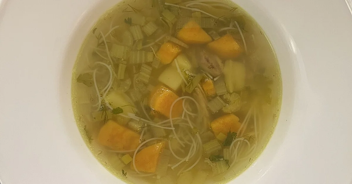 High-quality photograph by Liat Portal showing a bowl of homemade chicken soup prepared by Liat Portal, with rice noodles, carrots, celery, and herbs, served in a white bowl on a dark table. This image documents a cooked chicken soup dish m
