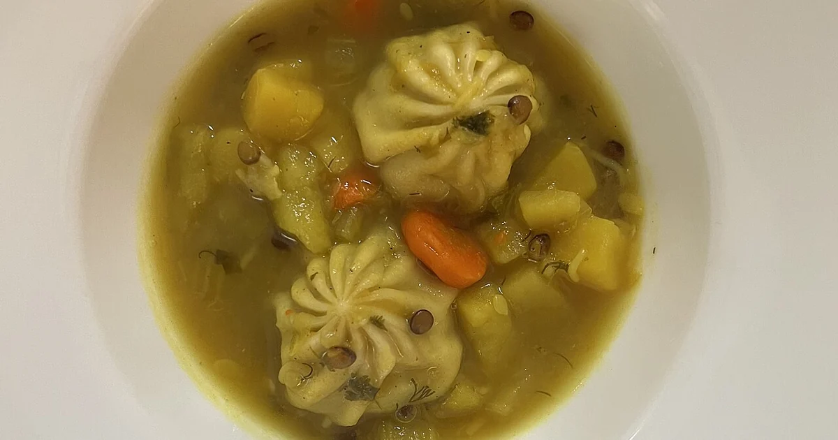 High-quality photograph by Liat Portal showing a bowl of homemade vegetable soup with carrots, potatoes, lentils, herbs, and chicken dumplings cooked in the broth. The soup was prepared by the photographer, with dumplings that were ready-ma