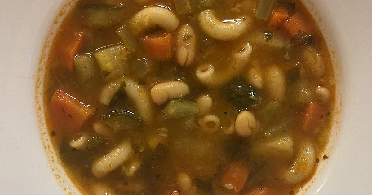 High-quality photograph by Liat Portal showing a bowl of minestrone soup cooked from scratch by the photographer. The soup contains beans, carrots, celery, herbs, and pasta in a tomato-based broth. This image documents everyday home cooking