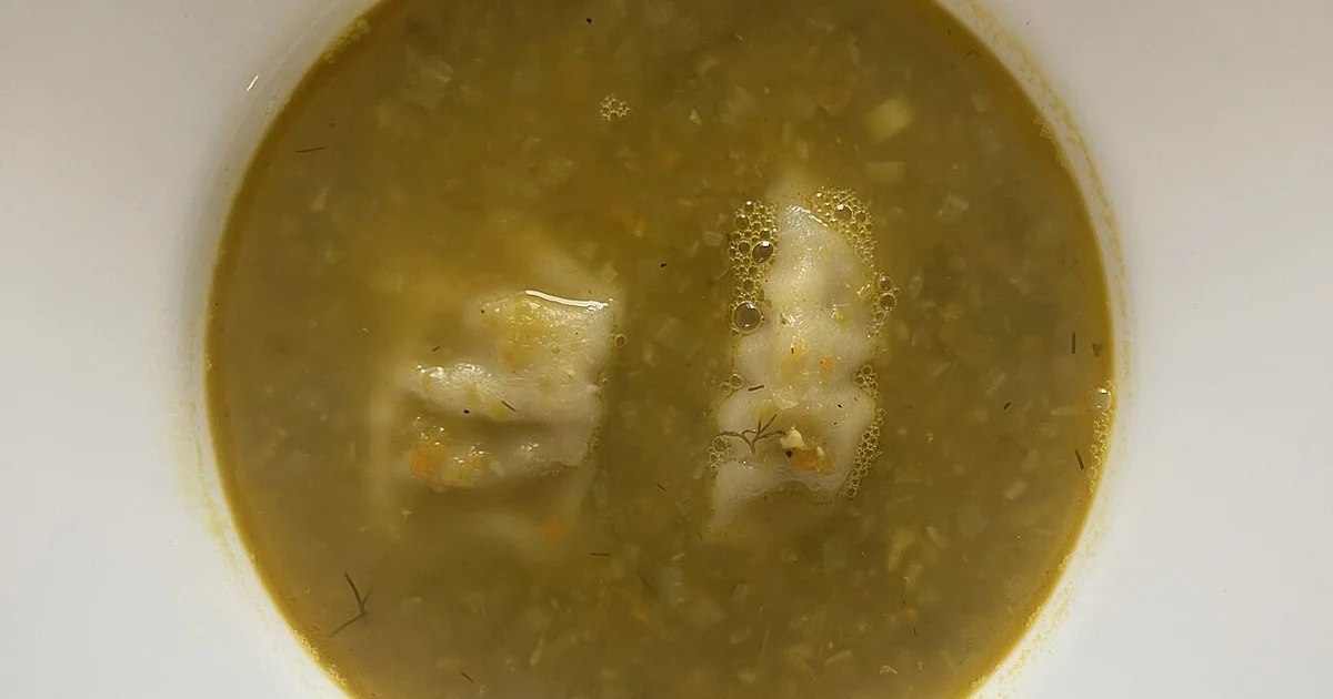 High-quality photograph by Liat Portal showing a bowl of homemade pea soup served in a white dish. The soup has a thick yellow-green broth with visible, finely chopped vegetables and two soft dumplings partially submerged in the liquid. The