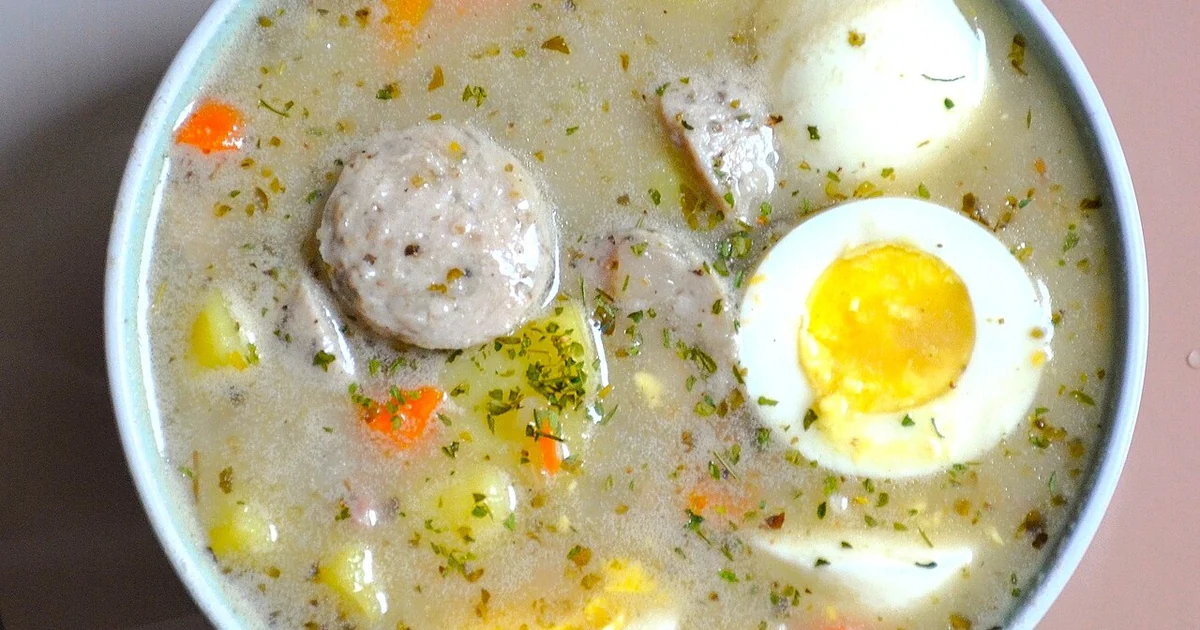 A top-down view (flat lay) of a sour rye soup prepared on a high-protein, long-simmered collagen broth base. The soup is served in a light blue bowl and contains traditional ingredients: sliced white sausage, halved hard-boiled eggs, potato