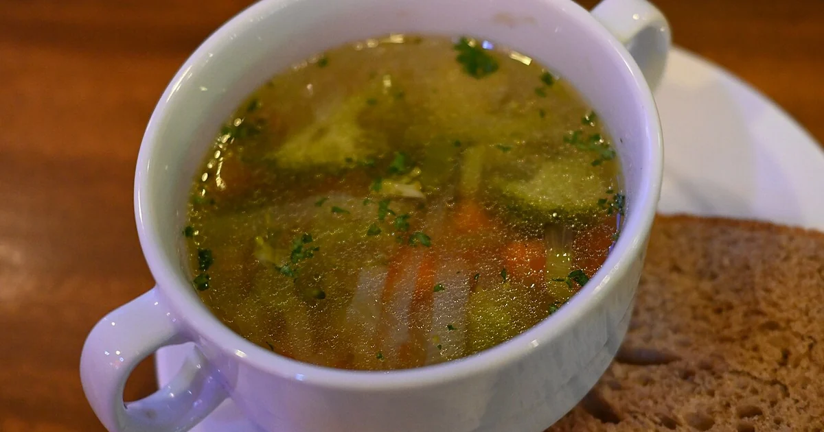 This photo shows a cup of clear broth soup in a white mug or cup with a handle. The soup appears to be a vegetable or chicken broth with visible ingredients floating in it, including what looks like small pieces of vegetables (possibly carr