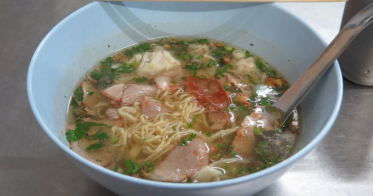 A bowl of noodle soup in Chiang Rai. The soup contains noodles, vegetables, sliced pork, and dumplings. It was purchased at an open-air restaurant. The soup is in a white bowl with a metal spoon and wooden chopsticks. Chiang Rai, Chiang Rai